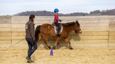In Aesch (8904, Kanton Zuerich) reitet ein Kind mit blauem Helm auf einem Pony in einem Sandviereck, waehrend eine Begleiterin das Pferd an der Longe fuehrt.