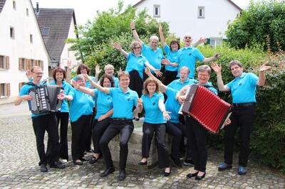 Eine Gruppe des Harmonika-Orchesters posiert mit ausgebreiteten Armen im Freien, zwei Personen halten Akkordeons, in Möhlin, 4313, Kanton Aargau.