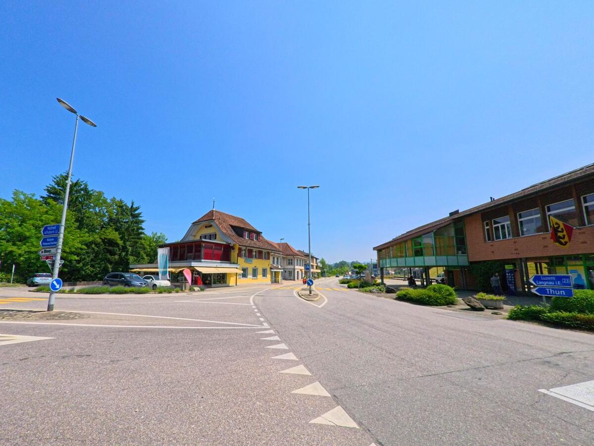 An einer belebten Kreuzung in Hasle bei Burgdorf (3415, Kanton Bern) treffen ein gelbes Gasthaus mit rotem Wintergarten und ein modernes Verwaltungsgebaeude aufeinander.