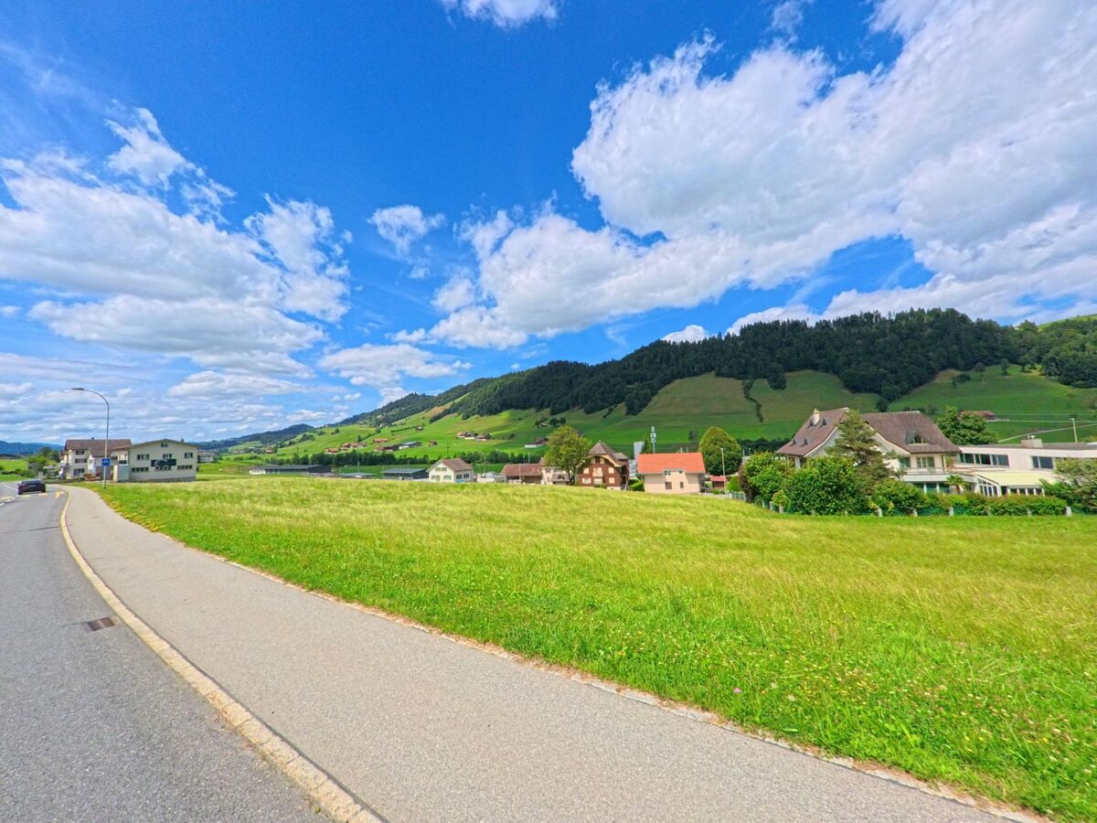 In Hasle (6166, Kanton Luzern) erstreckt sich eine offene Wiese mit Blick auf verstreute Häuser und bewaldete Hügel im Hintergrund.