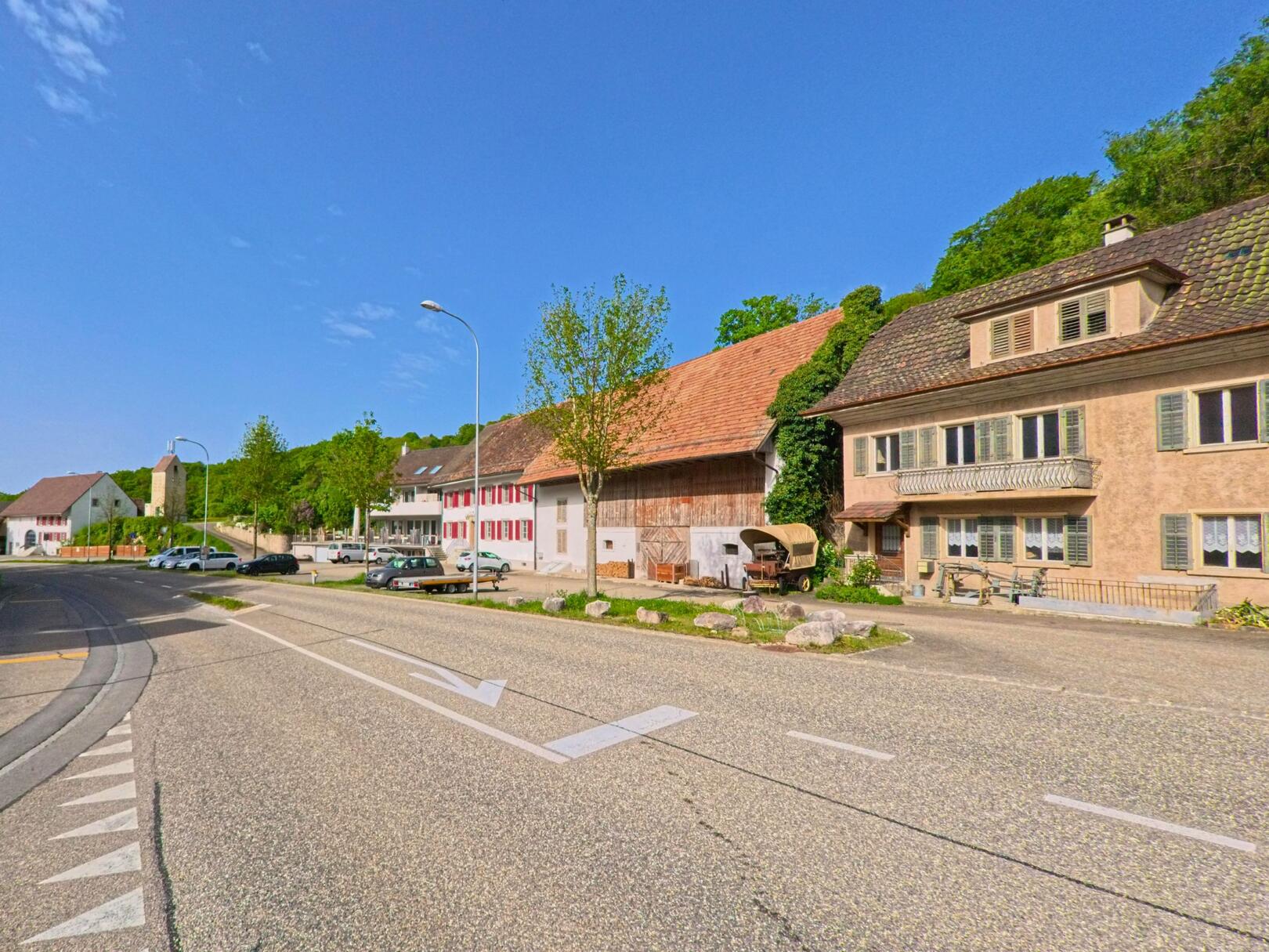 In Hauenstein-Ifenthal (4633, Kanton Solothurn) verläuft die Hauptstrasse an traditionellen Häusern und einem alten Wirtschaftsgebäude vorbei, mit Blick auf die Dorfkirche im Hintergrund.