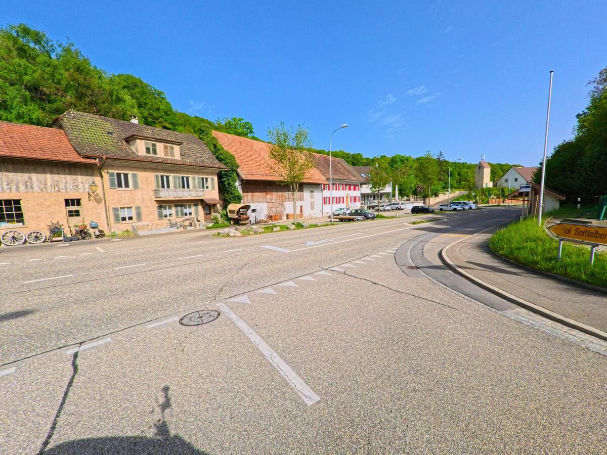 Die Hauptstrasse von Hauenstein-Ifenthal (4633, Kanton Solothurn) führt an historischen Gebäuden vorbei in Richtung der Dorfkirche, flankiert von Wäldern und einem Wegweiser zum Spittelberg.