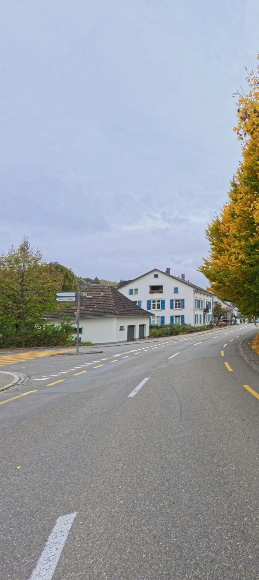 Eine breite Hauptstrasse mit gelben Markierungen fuehrt an einem weissen Gebaeude vorbei durch Hauptwil-Gottshaus (9213, Kanton Thurgau), flankiert von herbstlichen Baeumen.