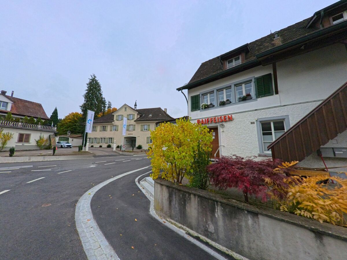 An einer Kurve neben einem Gebaeude mit Raiffeisen-Schriftzug zeigt sich ein Dorfzentrum von Hauptwil-Gottshaus (9213, Kanton Thurgau) mit herbstlicher Bepflanzung.