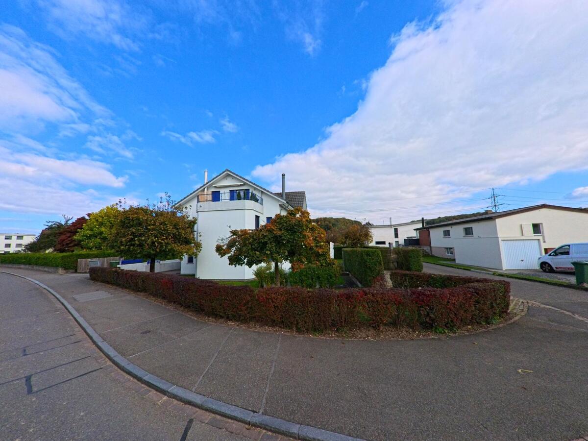 In einem ruhigen Wohnquartier von Hausen (5212, Kanton Aargau) steht ein helles Einfamilienhaus mit gepflegter Hecke und kleinen Baeumen an einer Strassenecke.