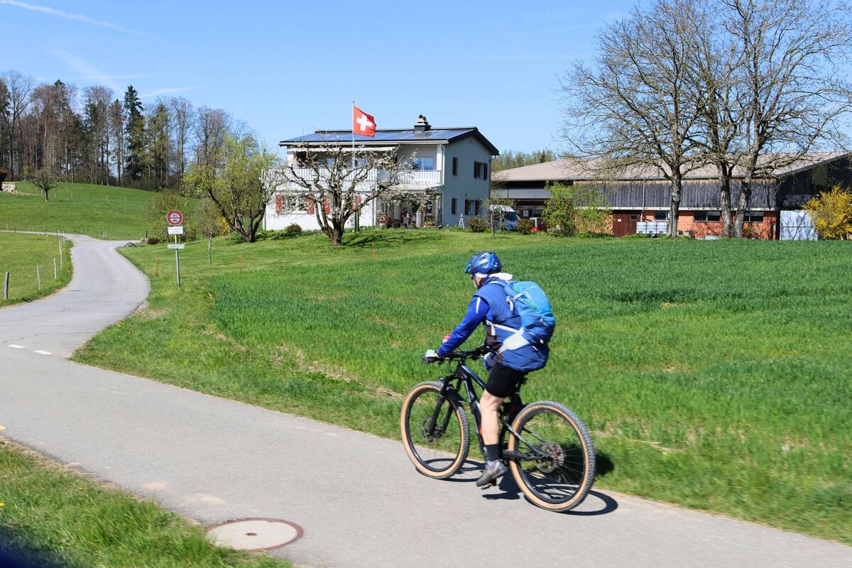 In Hausen am Albis (8915, Kanton Zürich) fährt ein Velofahrer auf einem asphaltierten Radweg durch eine grüne Wiesenlandschaft mit Bauernhof und Schweizer Flagge.