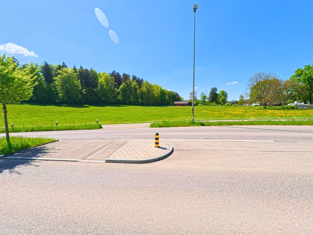 In Hausen am Albis (8915, Kanton Zürich) liegt eine Verkehrsinsel an einer Landstrasse mit Blick auf offene Wiesen und einen Waldrand im Hintergrund.