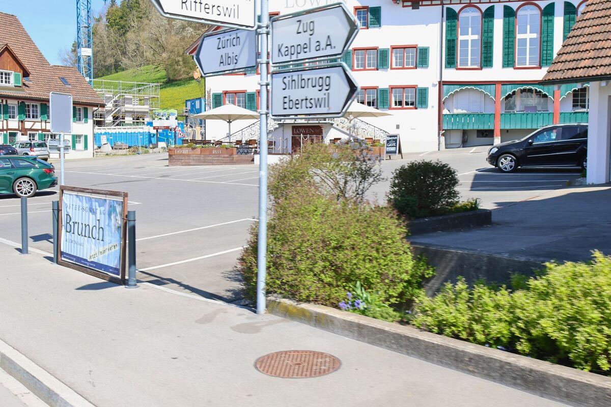 In Hausen am Albis (8915, Kanton Zürich) stehen mehrere Wegweiser an einer Strasse vor einem traditionellen Gasthof mit Parkplatz und Sonnenterrasse.