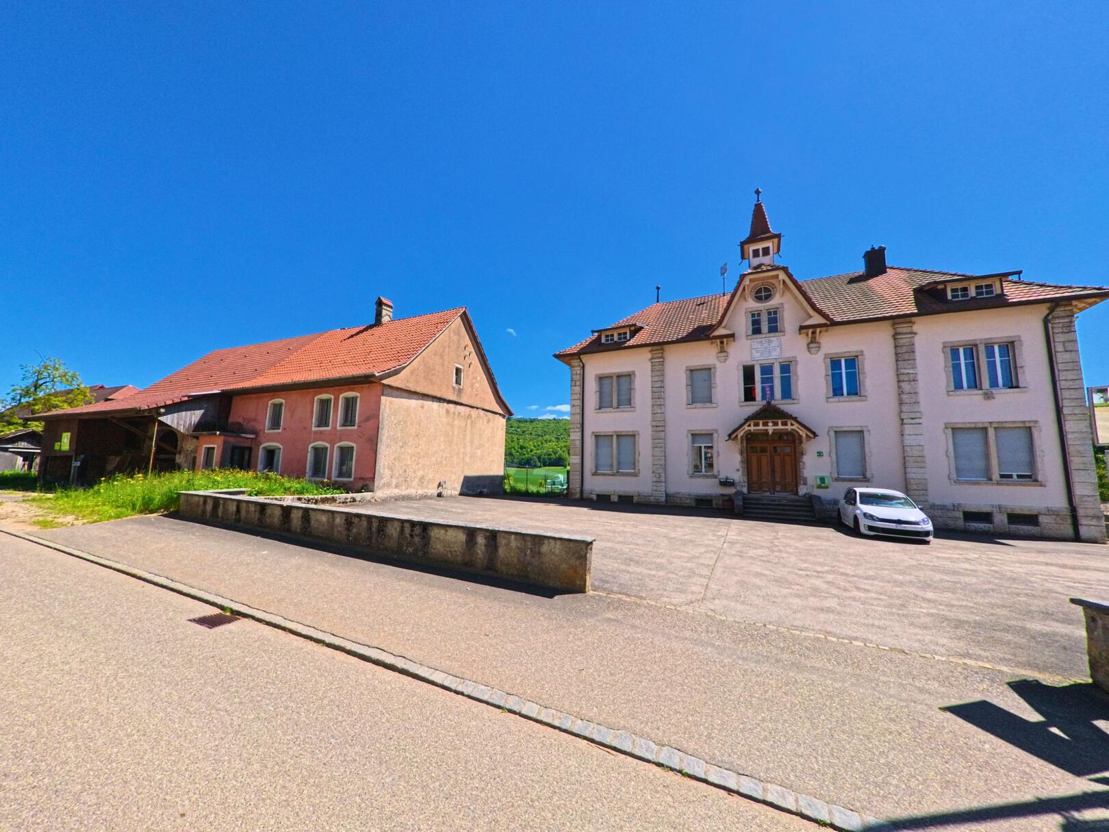 In Haute-Ajoie (2912, Kanton Jura) steht ein ehemaliges Schulhaus mit kleinem Türmchen und grossem Vorplatz neben einem älteren, rötlich verputzten Gebäude.