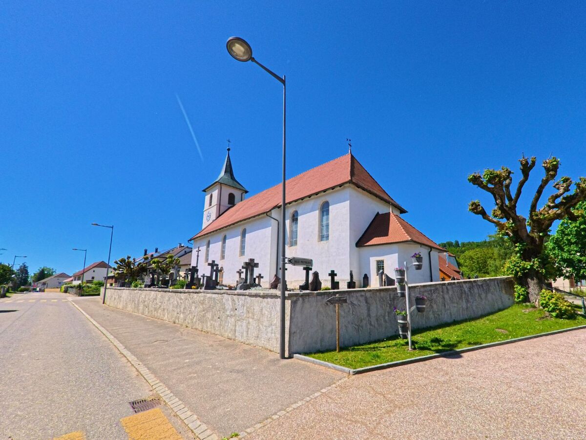 Die Kirche von Haute-Ajoie (2914, Kanton Jura) mit ihrem roten Satteldach und angrenzendem Friedhof liegt erhöht an einer ruhigen Strasse.