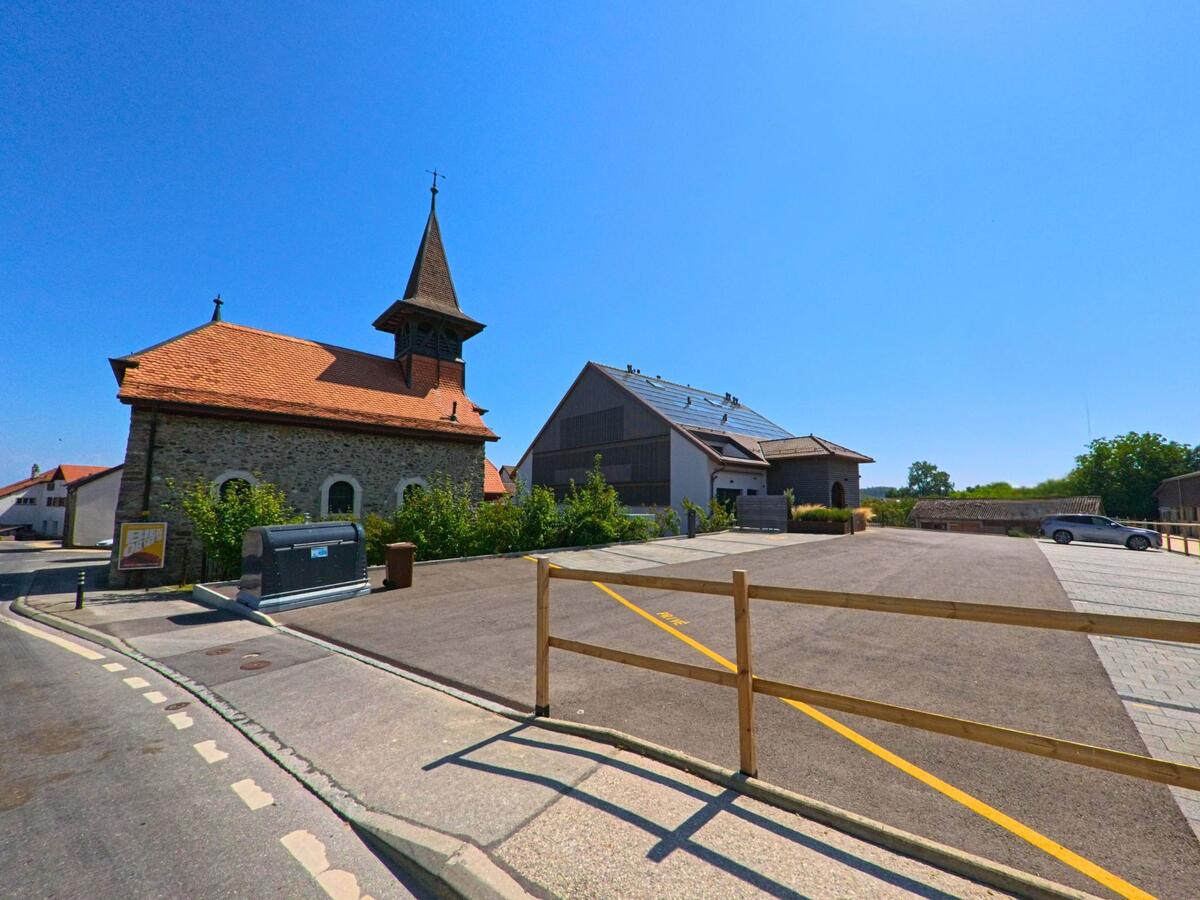 Die kleine Kirche mit ihrem Turm steht im Zentrum von Hautemorges (1141, Kanton Waadt) neben modernen Gebäuden und einem Parkplatz.