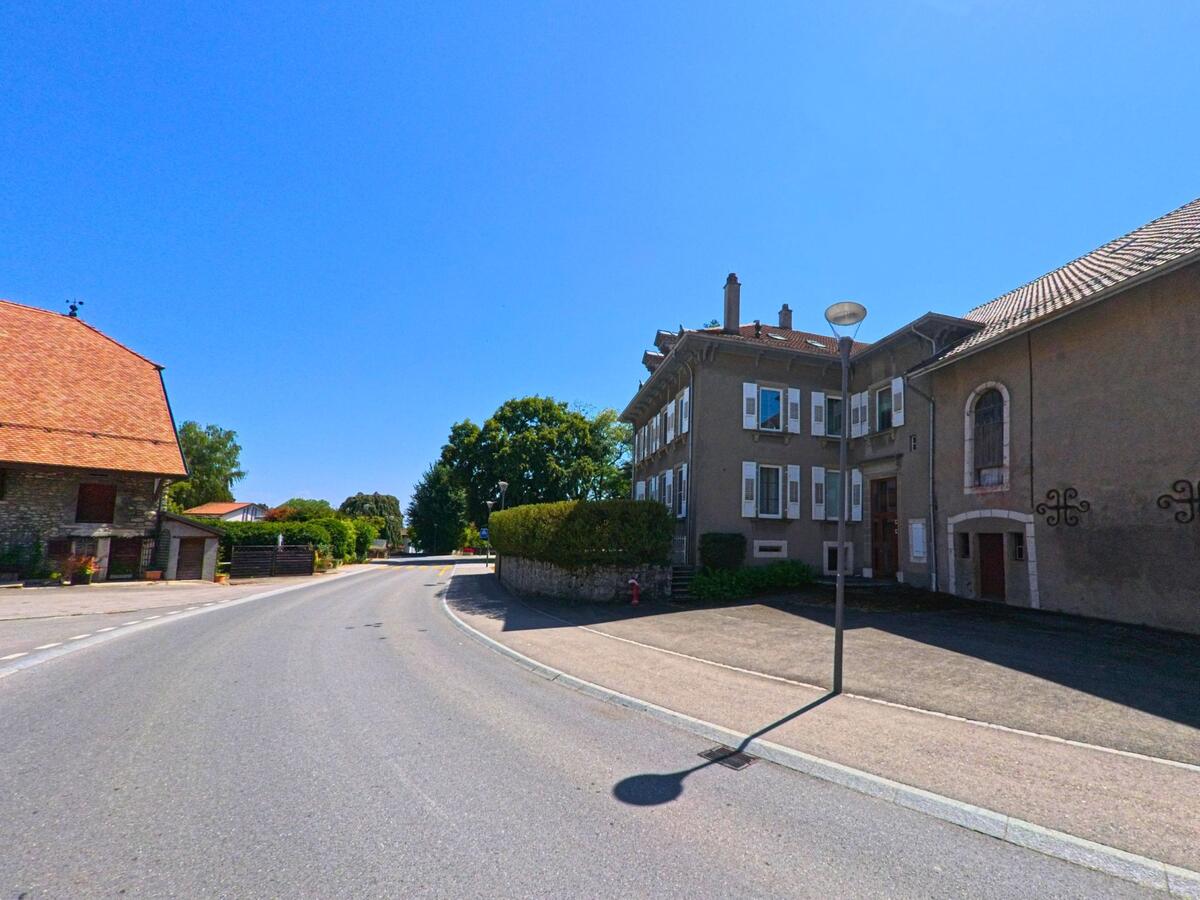 An einer breiten Strasse in Hautemorges (1143, Kanton Waadt) stehen traditionelle Wohnhaeuser mit Fensterlaeden und angrenzenden Steinmauern.