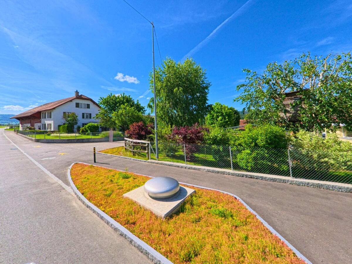 In Haute-Sorne (2853, Kanton Jura) liegt eine bepflanzte Verkehrsinsel mit moderner Leuchte vor Wohnhäusern und gepflegten Gärten am Strassenrand.