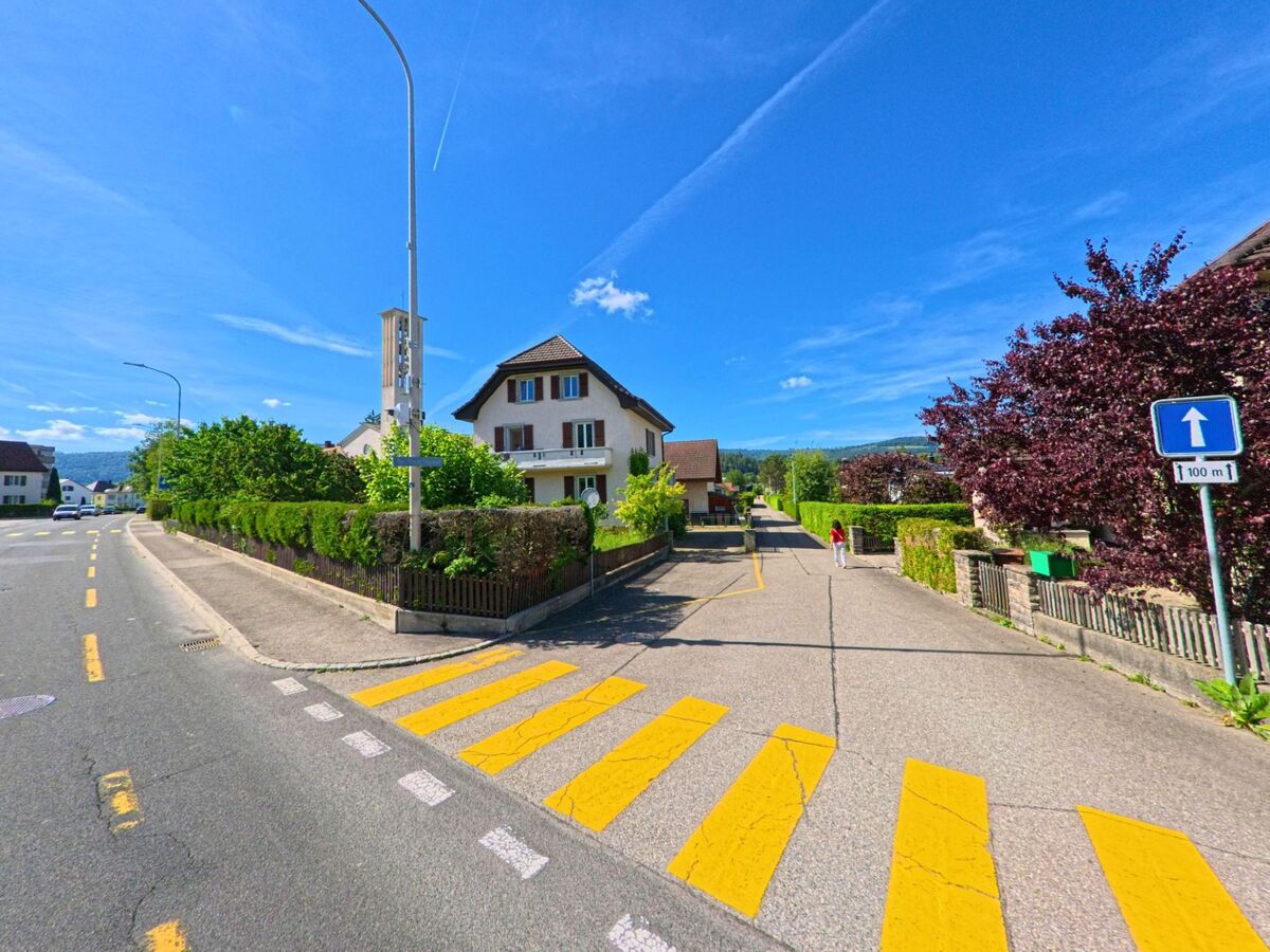 In Haute-Sorne (2854, Kanton Jura) beginnt eine Einbahnstrasse mit gelbem Fussgängerstreifen vor einem Wohnhaus mit roten Fensterläden und gepflegter Hecke.