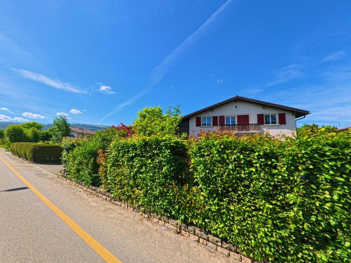 In Haute-Sorne (2854, Kanton Jura) steht ein Einfamilienhaus mit roten Fensterläden hinter einer dichten, gepflegten Hecke an einer sonnigen Quartierstrasse.
