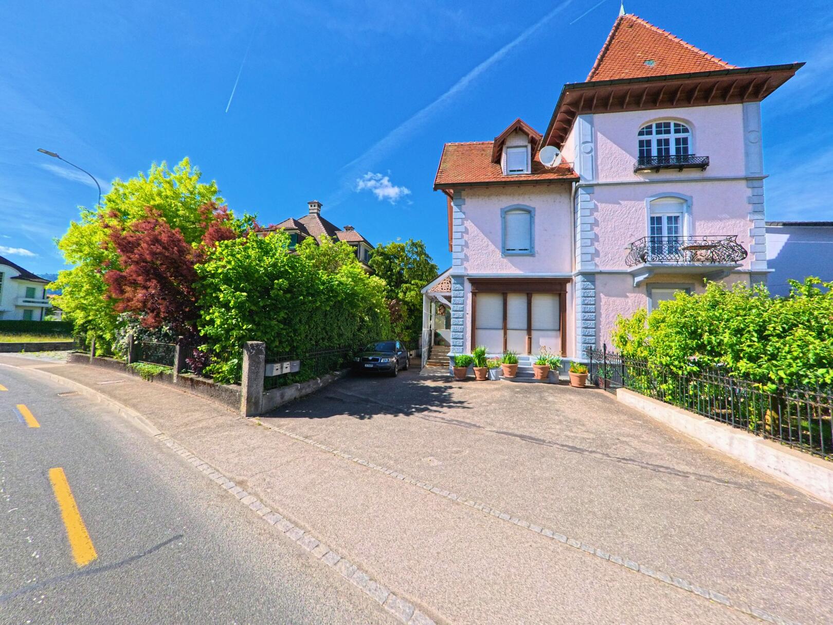 Ein herrschaftliches Wohnhaus mit Türmchen und Balkonen steht in Haute-Sorne (2854, Kanton Jura) direkt an einer leicht gebogenen Strasse, umgeben von Hecken und Einfahrt.
