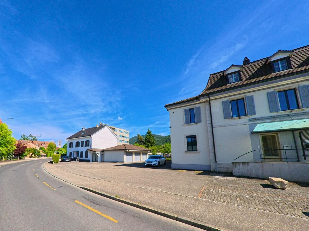 Entlang einer leicht geschwungenen Strasse in Haute-Sorne (2854, Kanton Jura) stehen helle Wohnhäuser mit gepflegten Vorplätzen und Blick auf bewaldete Hügel.