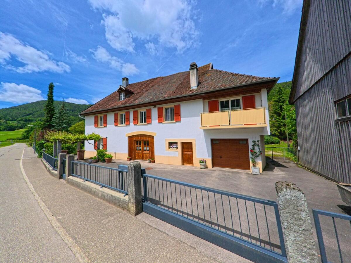 Ein gepflegtes Wohnhaus mit roten Fensterläden und Holztor steht in Haute-Sorne (2864, Kanton Jura) an einer schmalen Strasse mit Blick auf grüne Hügel.