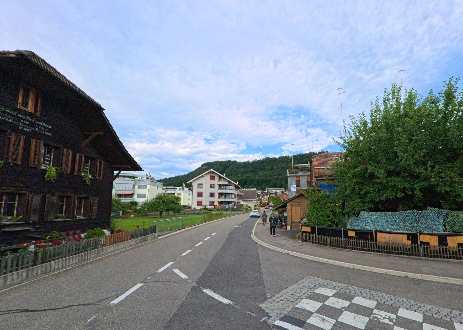 In Heimberg (3627, Kanton Bern) verläuft eine Hauptstrasse an traditionellen und modernen Wohnhäusern vorbei in Richtung eines bewaldeten Hügels.