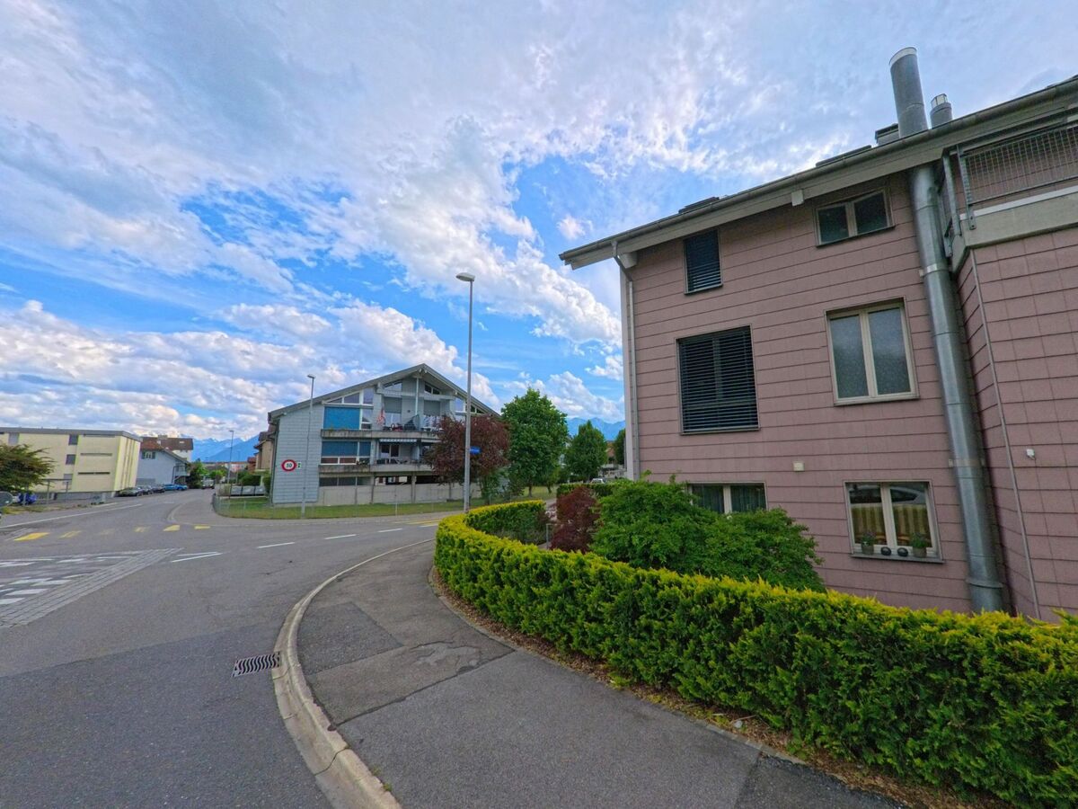 In Heimberg (3627, Kanton Bern) säumen Mehrfamilienhäuser mit gepflegter Bepflanzung eine leicht geschwungene Quartierstrasse mit Blick in Richtung Alpen.