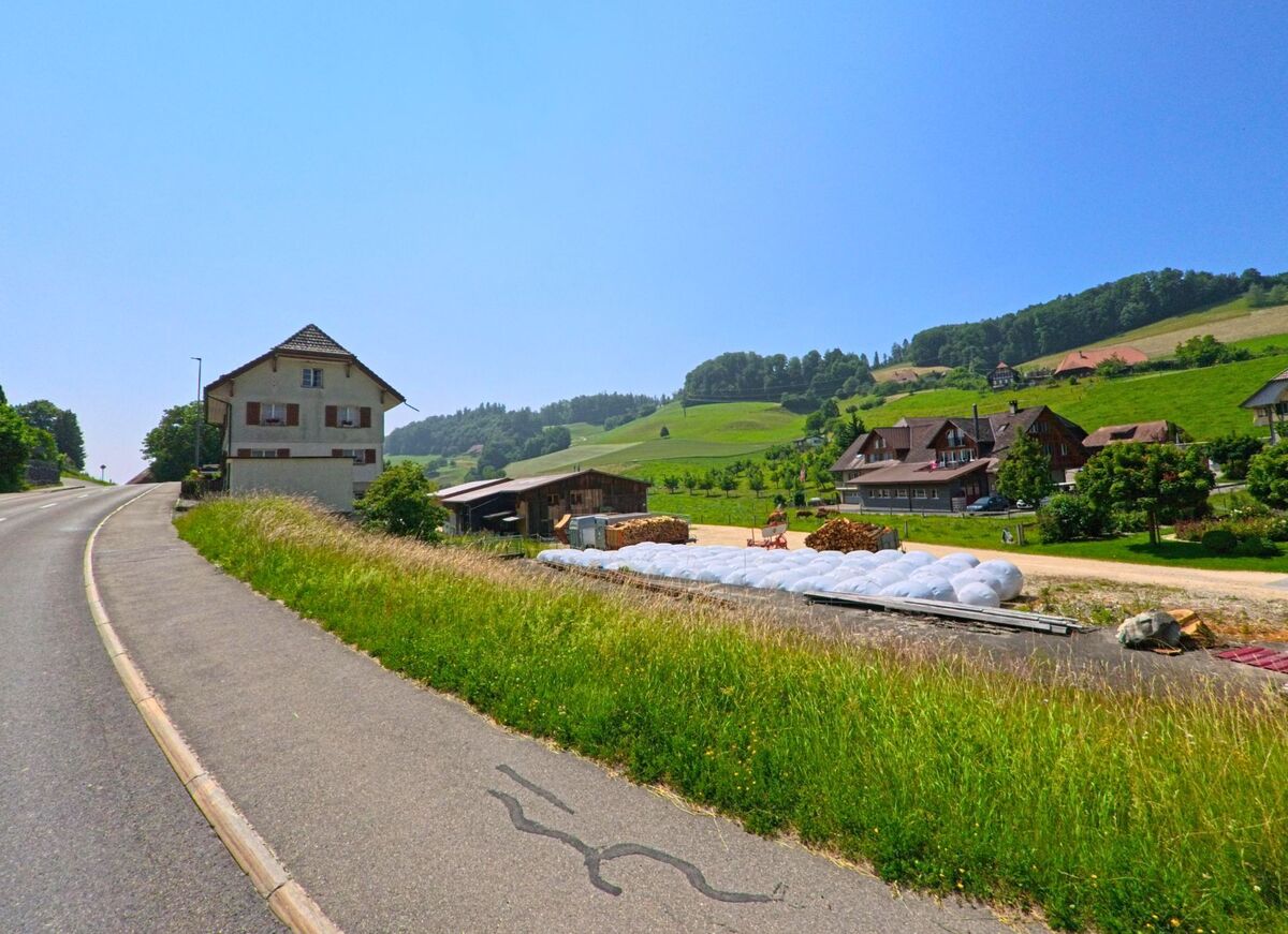 Entlang einer leicht ansteigenden Strasse in Heimiswil (3412 im Kanton Bern) liegen Bauernhöfe und Wohnhäuser eingebettet in die grüne Hügellandschaft, mit Siloballen im Vordergrund.