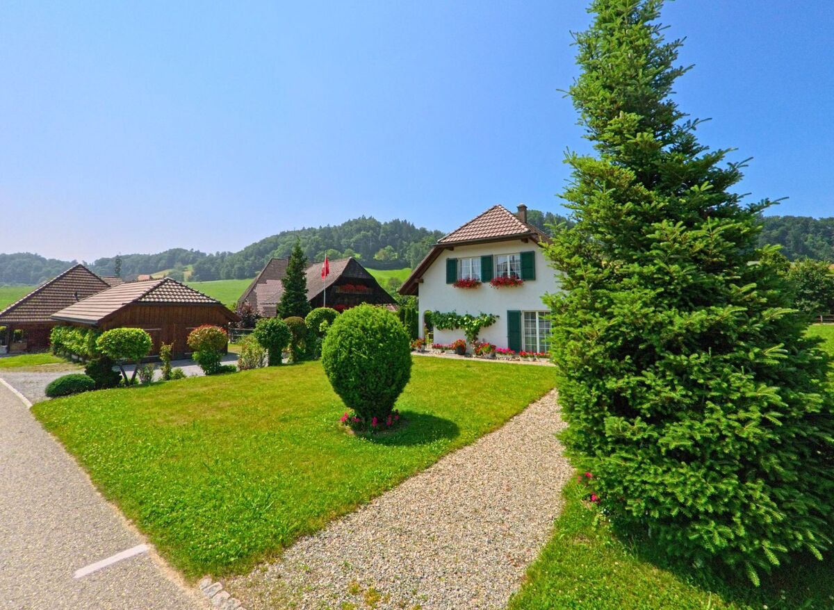 In Heimiswil (3412, Kanton Bern) steht ein gepflegtes Einfamilienhaus mit Blumenschmuck und Gartenanlage inmitten der ländlichen Hügellandschaft mit traditionellen Gebäuden im Hintergrund.