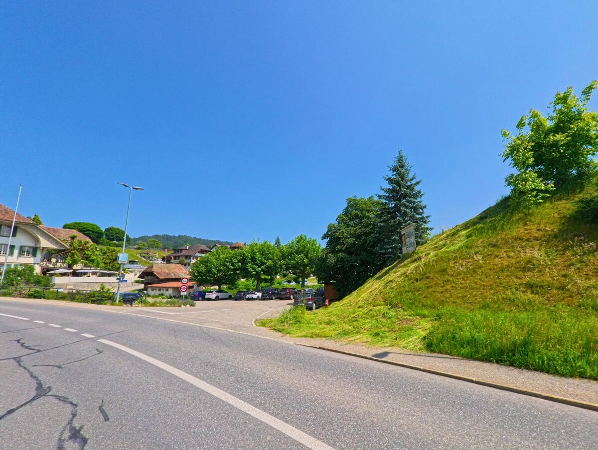 Eine sanft geschwungene Hauptstrasse führt durch Heimiswil (3412 im Kanton Bern), vorbei an einem Parkplatz und mehreren Wohnhäusern, während ein bewachsener Hügel das Strassenbild prägt.
