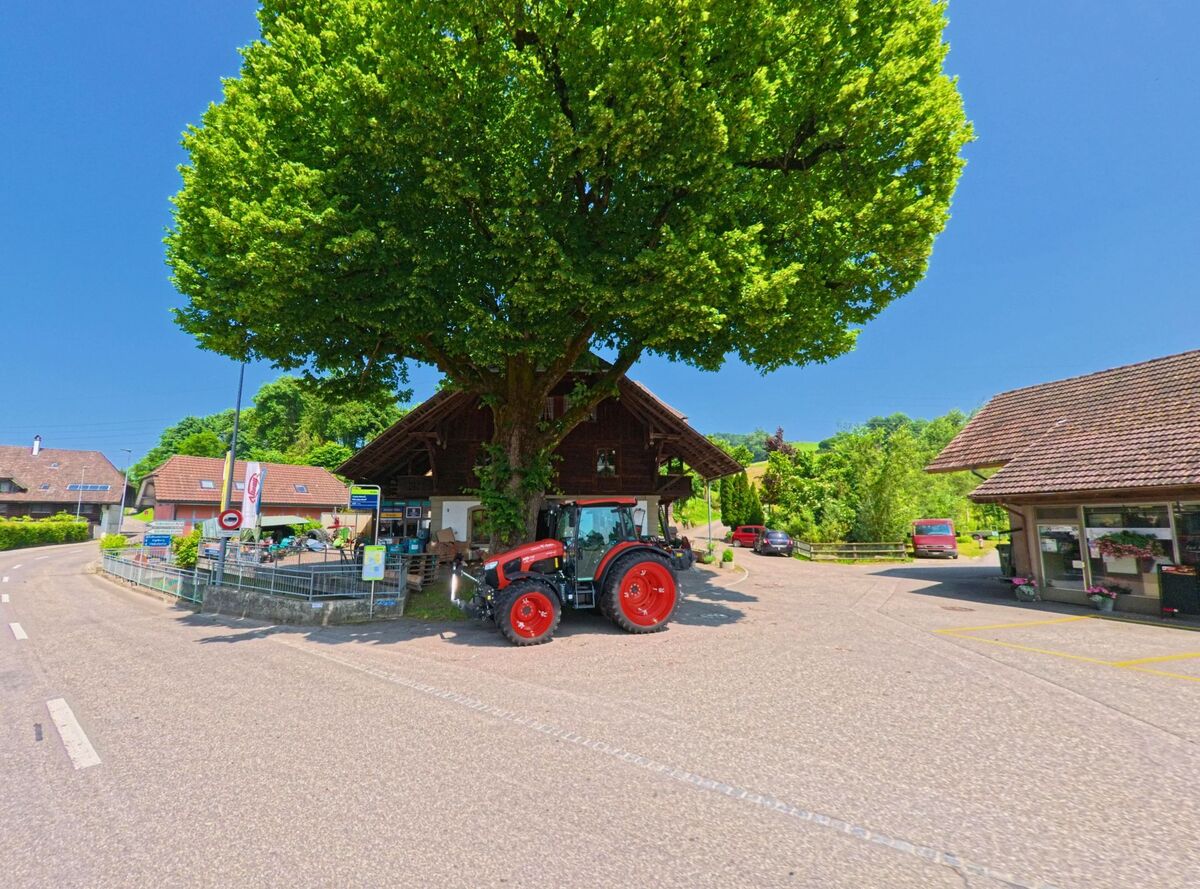 Unter einer ausladenden Linde steht ein roter Traktor vor einem traditionellen Gebäude im Zentrum von Heimiswil (3412 im Kanton Bern), das als Treffpunkt und Verkaufsstelle dient.