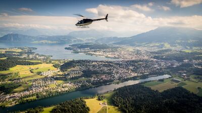 Ein Helikopter fliegt über die Landschaft bei Wohlenschwil (5512, Kanton Aargau) mit Blick auf grüne Felder, Wälder und den See im Hintergrund.