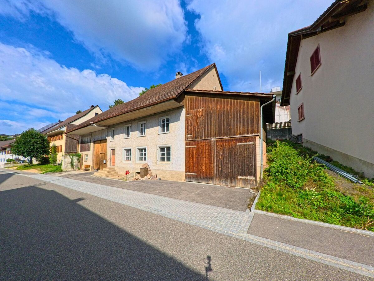 Ein traditionelles Bauernhaus mit Holztor und hellem Putz steht in Hellikon (4316, Kanton Aargau) entlang einer ruhigen Dorfstrasse bei Sonnenschein.