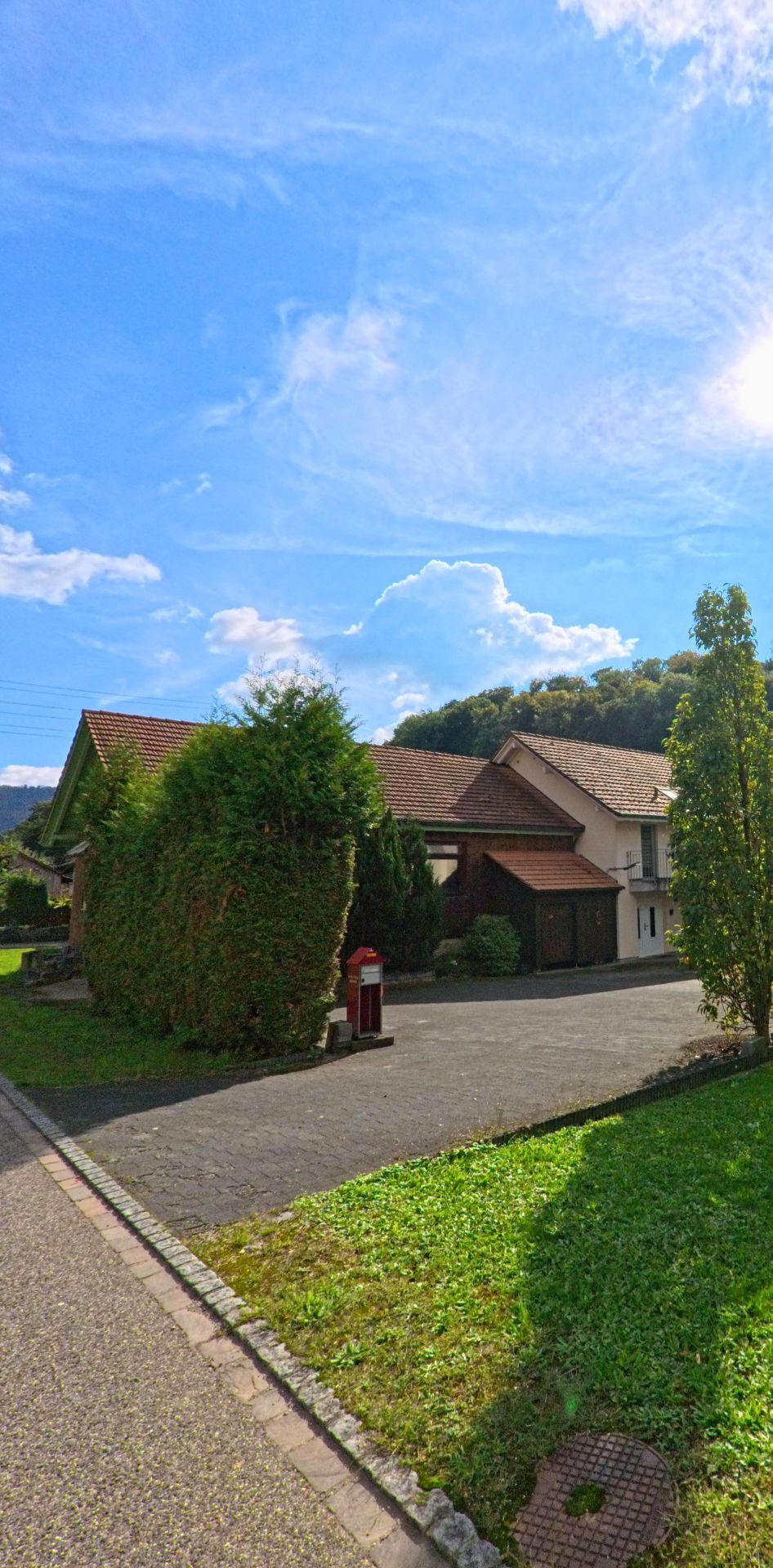 Ein Einfamilienhaus mit Ziegeldach und dichten Hecken steht in Hellikon (4316, Kanton Aargau) an einer ruhigen Nebenstrasse bei sonnigem Wetter.