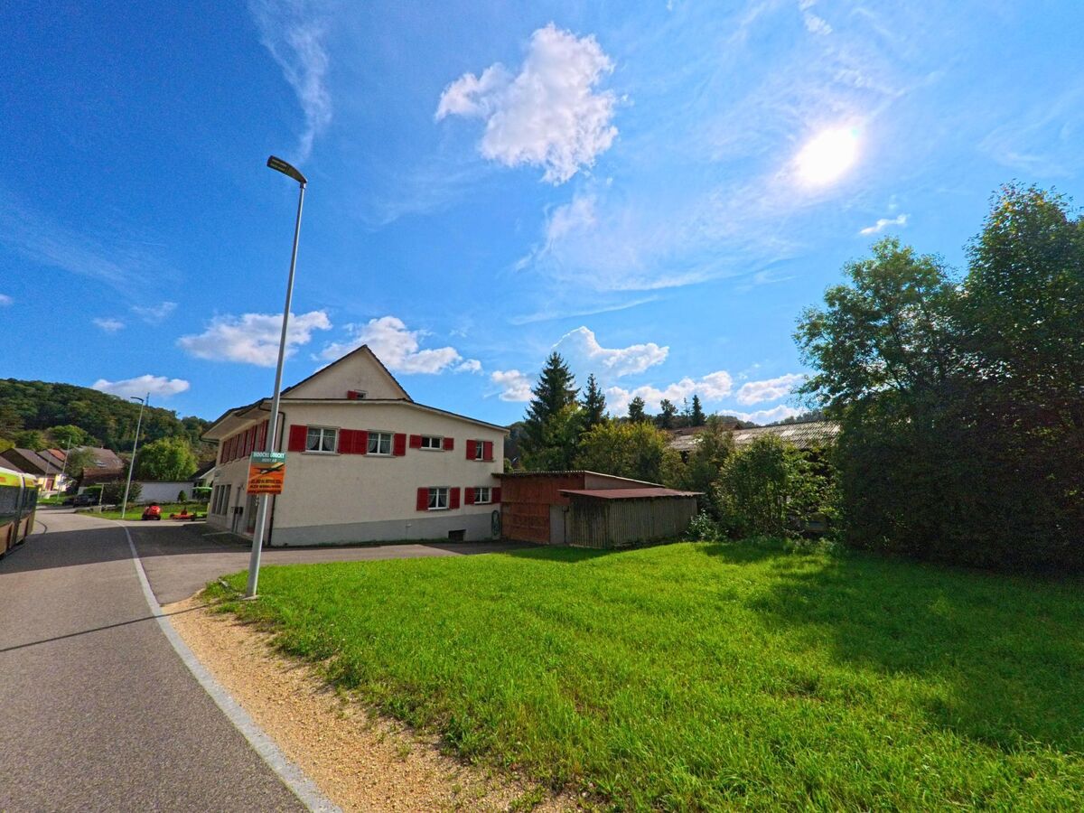 Ein Haus mit roten Fensterlaeden steht in Hellikon (4316, Kanton Aargau) an einer ruhigen Strasse neben einer gruene Wiese unter strahlender Sonne.
