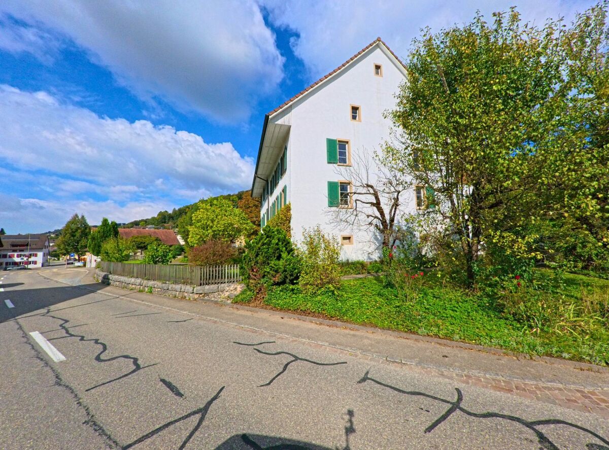 An der Hauptstrasse von Hellikon (4316, Kanton Aargau) steht ein weisses Haus mit gruenen Fensterlaeden, umgeben von Baeumen und einem kleinen Garten.
