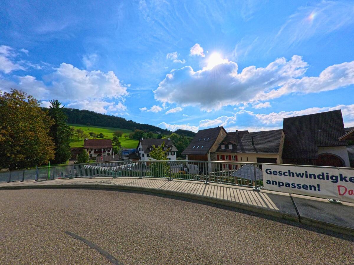 Über dem sonnigen Dorfrand von Hemmiken (4465, Kanton Basel-Landschaft) spannt sich der Blick von einer leicht erhöhten Strasse über Häuser und weite, grüne Hügel.