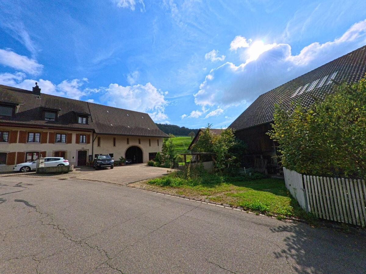 Hinter einem grossen Bauernhaus in Hemmiken (4465, Kanton Basel-Landschaft) öffnet sich der Blick auf die sonnigen Felder, während gegenüber eine Scheune teils im Schatten liegt.
