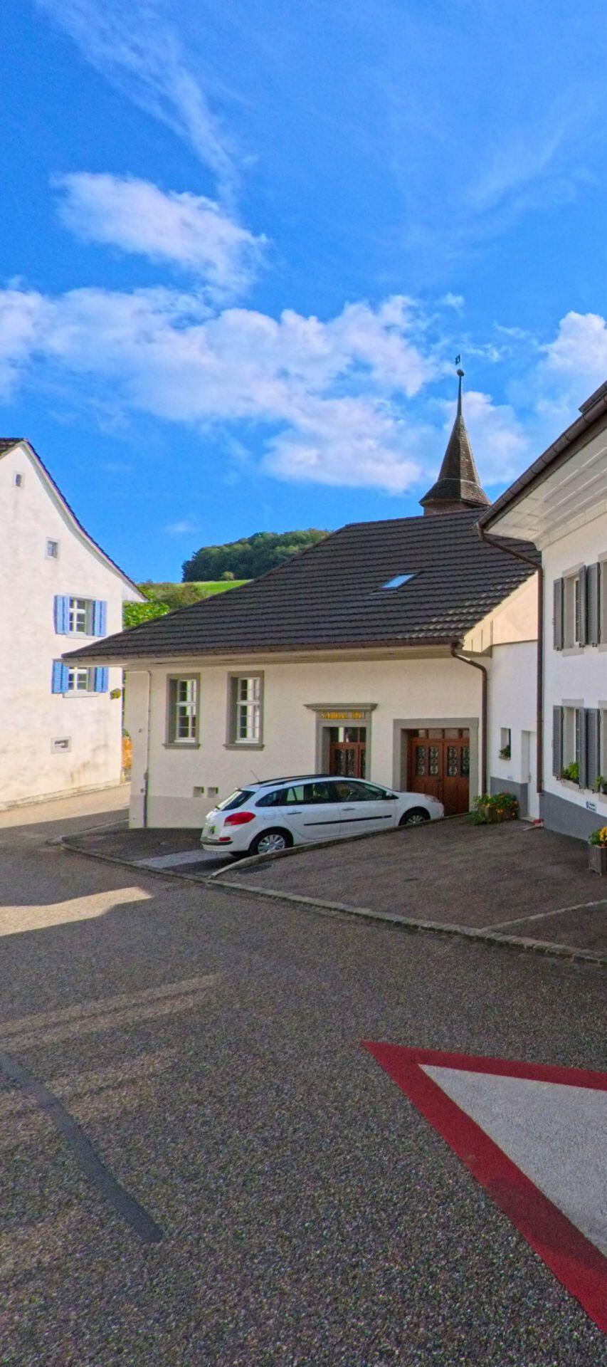 Vor dem Gebäude mit dem spitzen Turm in Hemmiken (4465, Kanton Basel-Landschaft) verläuft eine ruhige Strasse mit einem geparkten Auto und Blick auf die umliegenden Hügel.