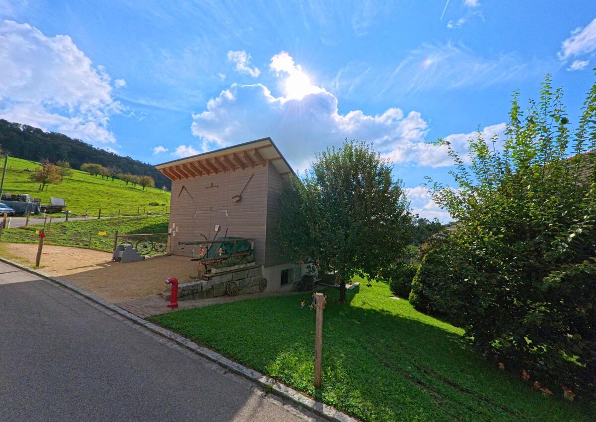 Hinter einem kleinen Gebäude mit alten landwirtschaftlichen Geräten in Hemmiken (4465, Kanton Basel-Landschaft) breiten sich sonnige Weiden vor dem bewaldeten Hang aus.