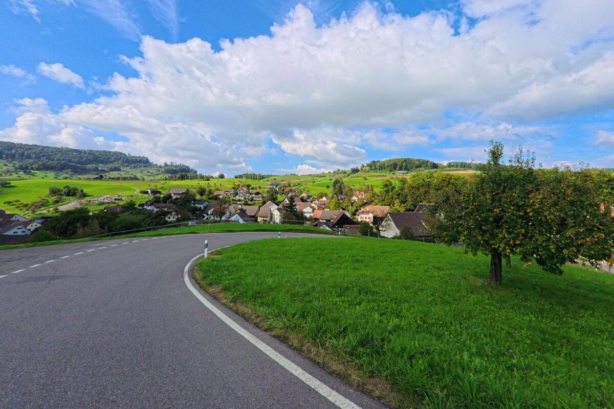 Von einer geschwungenen Strasse oberhalb von Hemmiken (4465, Kanton Basel-Landschaft) öffnet sich der Blick über das Dorf und die weitläufigen, grünen Hügel im Hintergrund.