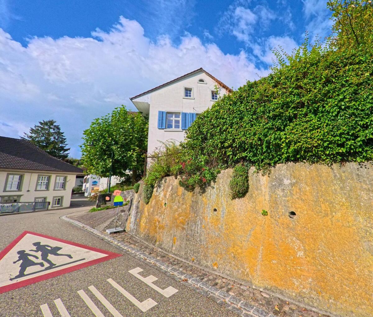 An einer Schulwegmarkierung in Hemmiken (4465, Kanton Basel-Landschaft) verläuft die Strasse entlang einer hohen, bewachsenen Mauer unterhalb eines Hauses mit blauen Fensterläden.