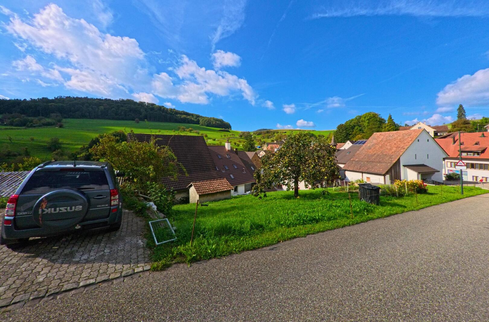 Über den Dächern von Hemmiken (4465, Kanton Basel-Landschaft) zeigt das Bild eine ruhige Aussicht auf die Häuserzeilen vor den grünen Hügeln und dem klaren Himmel.
