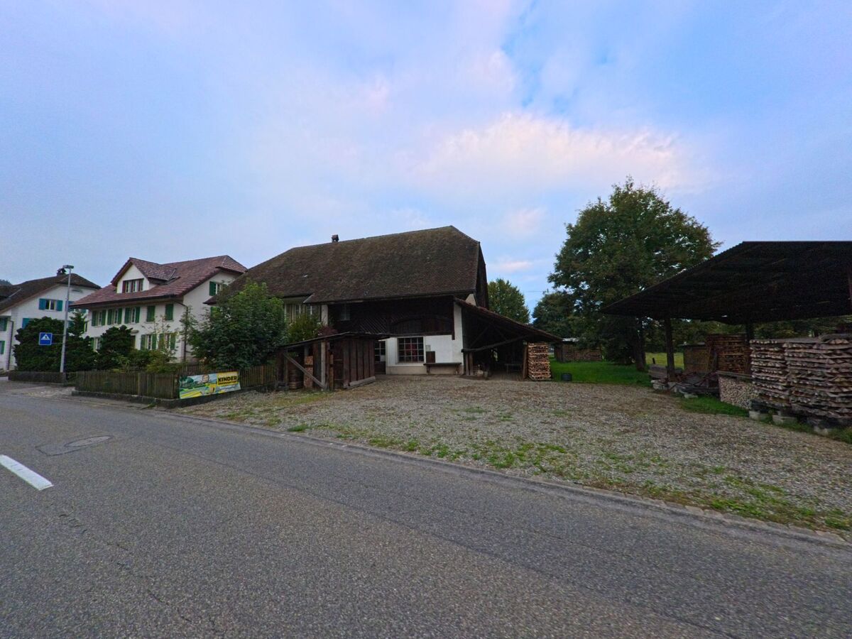 Ein Bauernhof mit grossem Scheunendach und gestapeltem Brennholz steht in Hendschiken (5604, Kanton Aargau) neben einer Landstrasse.