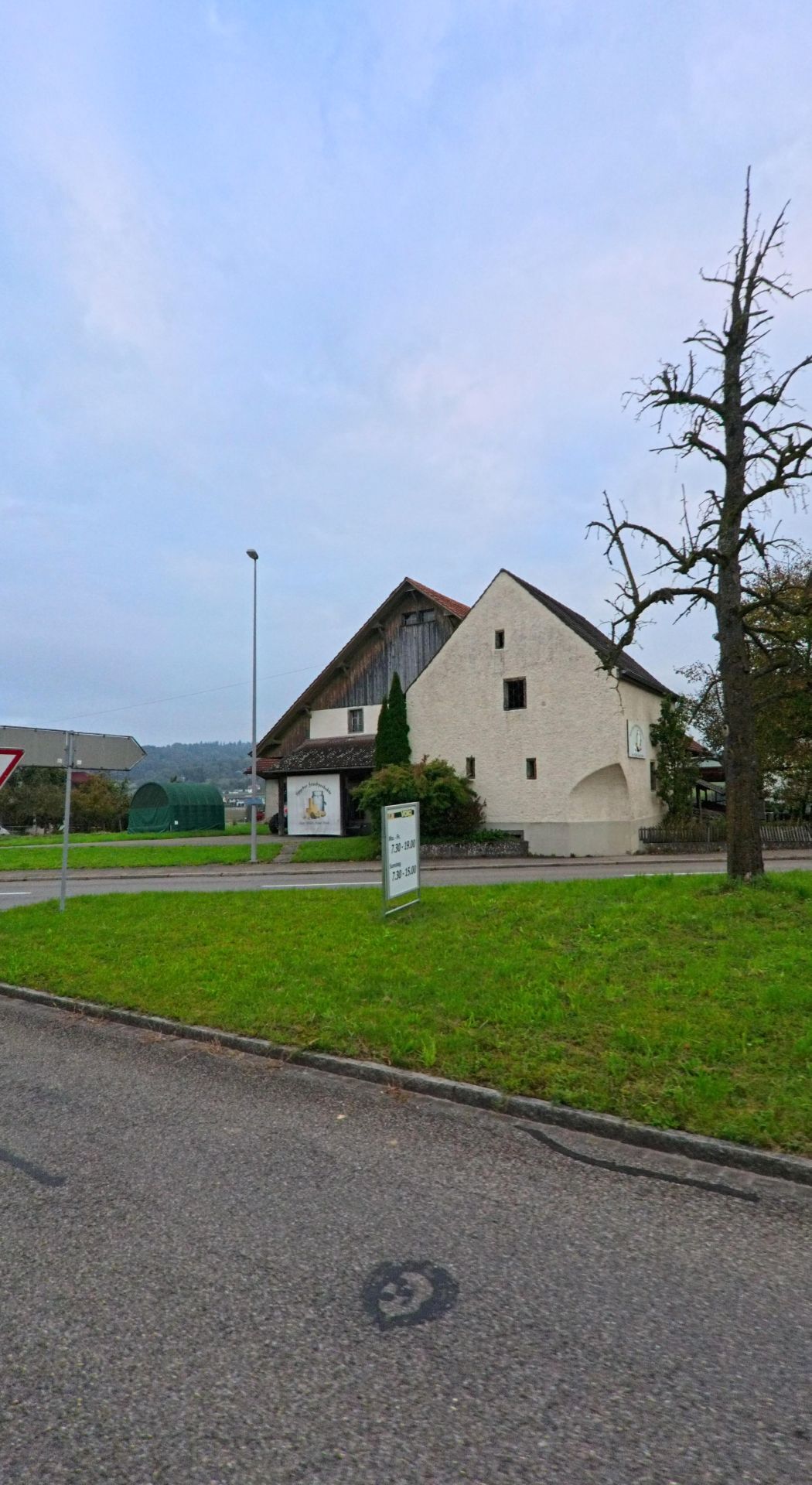 Ein traditionelles Gebaeude mit heller Fassade und Scheunendach steht in Hendschiken (5604, Kanton Aargau) an einer Strassenkreuzung mit gruener Wiese im Vordergrund.
