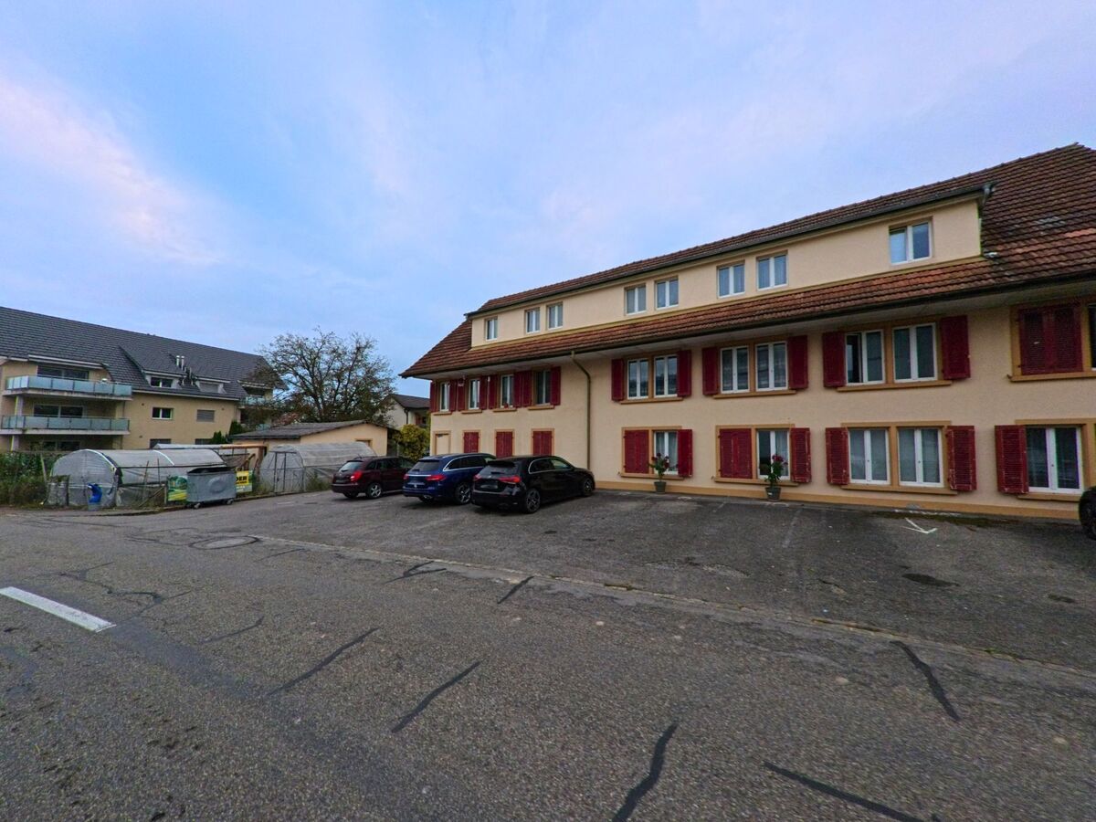 Vor einem laenglichen Wohnhaus mit roten Fensterlaeden in Hendschiken (5604, Kanton Aargau) befinden sich Parkplaetze und kleine Gewaechshaeuser.