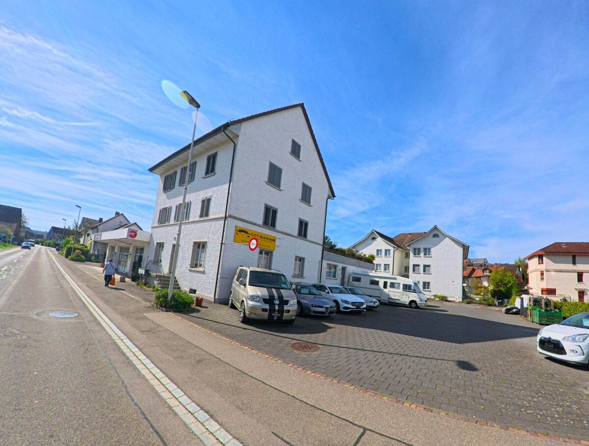 In Henggart im Kanton Zürich mit der PLZ 8444 verläuft eine Hauptstrasse entlang eines Garagenbetriebs mit angrenzendem Parkplatz und Wohnhäusern im Hintergrund.