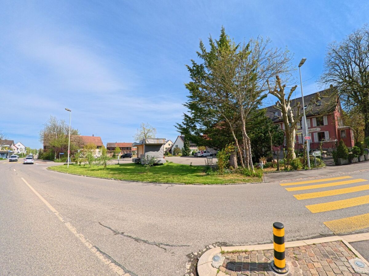 Ein Kreisverkehr in Henggart, PLZ 8444 im Kanton Zürich, verbindet mehrere Quartierstrassen, flankiert von Bäumen, Zebrastreifen und einem historischen Gebäude mit roter Fassade.