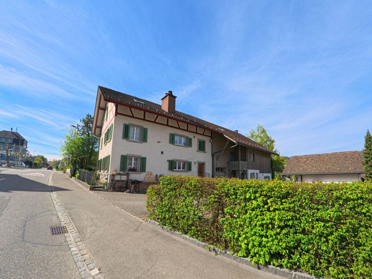 Ein traditionelles Wohnhaus mit grünen Fensterläden und angebautem Scheunentrakt steht in Henggart im Kanton Zürich, PLZ 8444, entlang einer sanft geschwungenen Strasse.