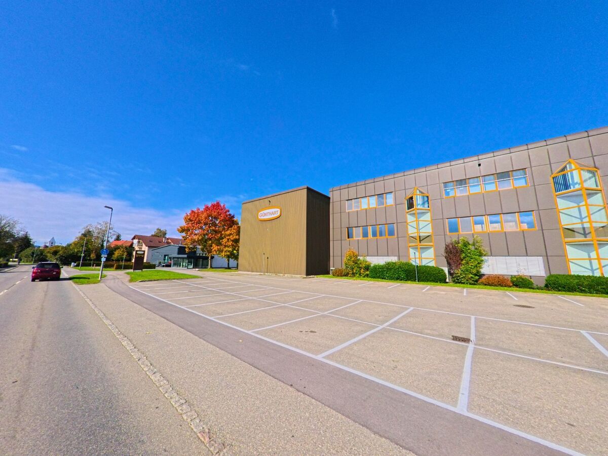 Ein modernes Gewerbegebäude mit grossem Parkplatz liegt in Herdern (8535, Kanton Thurgau) an einer breiten Strasse unter blauem Himmel.