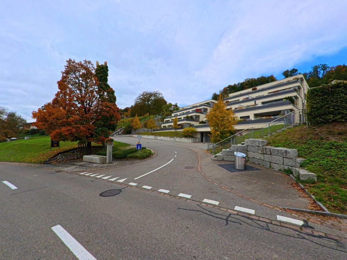 An einer Kurve in Herdern (8535, Kanton Thurgau) führen Strasse und Treppen an einem Hang mit modernen Terrassenwohnungen und herbstlichen Bäumen vorbei.