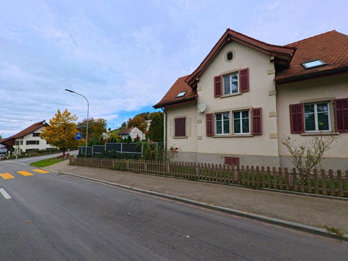 An einer Strasse mit Fussgängerstreifen steht in Herdern (8535, Kanton Thurgau) ein Wohnhaus mit roten Fensterläden hinter einem niedrigen Gartenzaun.