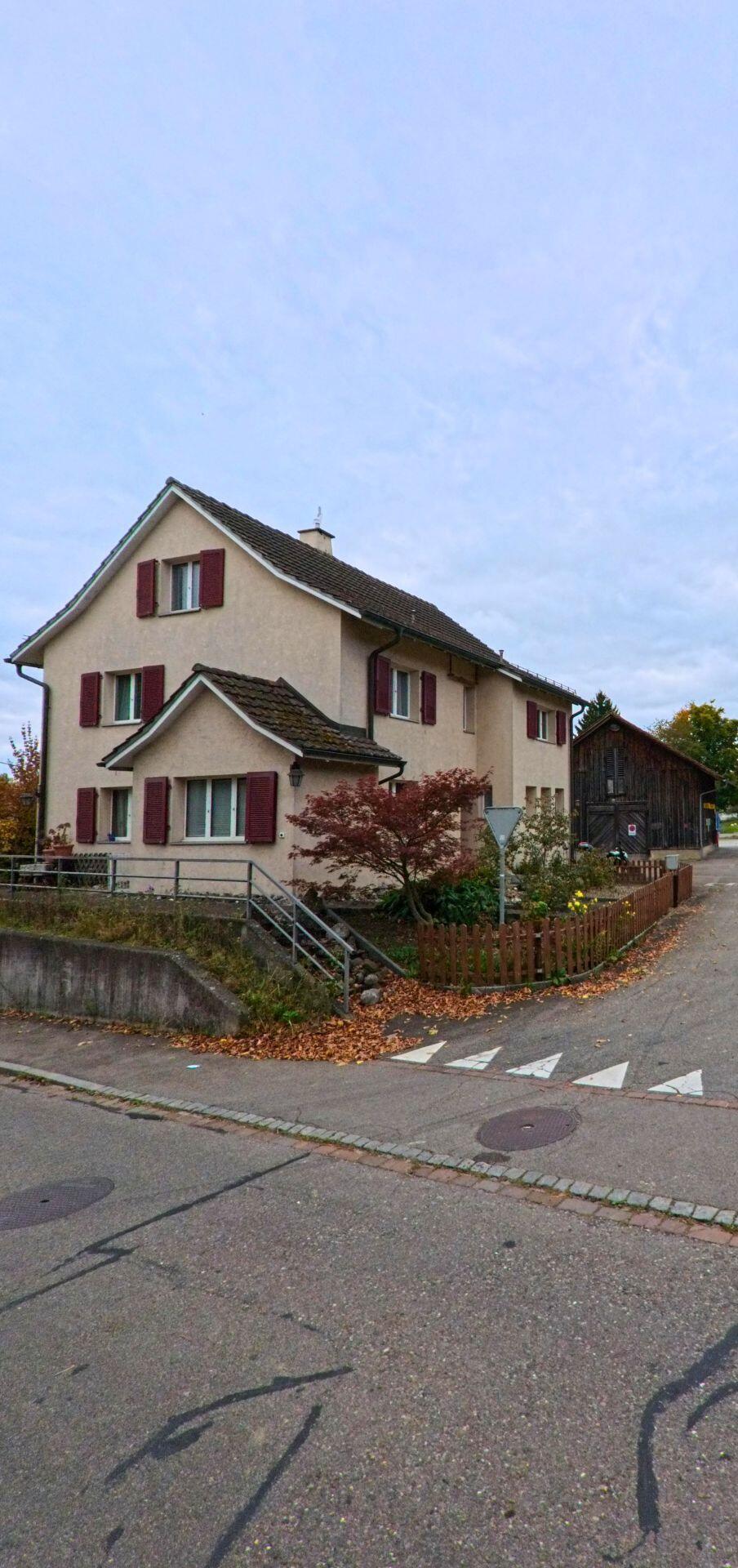 Ein Wohnhaus mit roten Fensterläden steht in Herdern (8535, Kanton Thurgau) an einer kleinen Kreuzung neben einem Holzschopf und einem herbstlichen Garten.